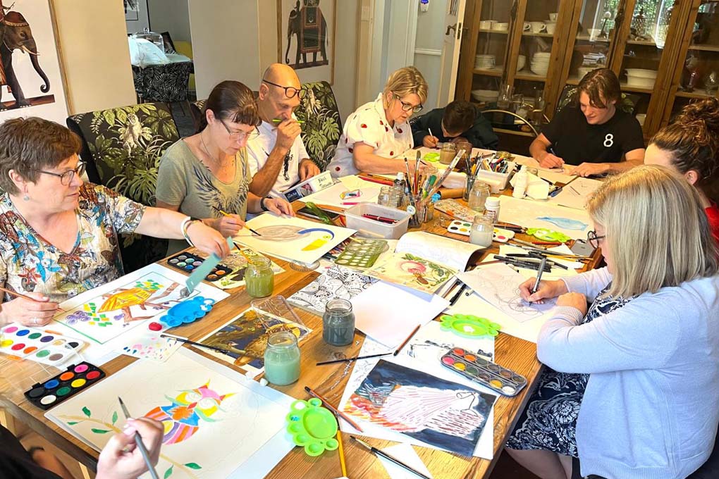 Art Artist working on a painting at a wooden table in a sunlit art studio filled with canvases, brushes, and colourful paint splashes.